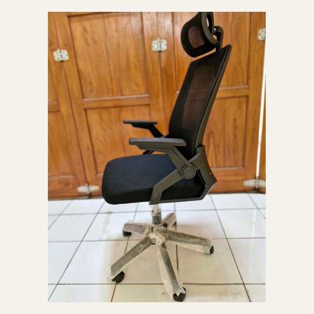 Kursi kantor hitam tampak serong dengan headrest miring, sandaran jaring, dan kaki beroda terbungkus plastik.
