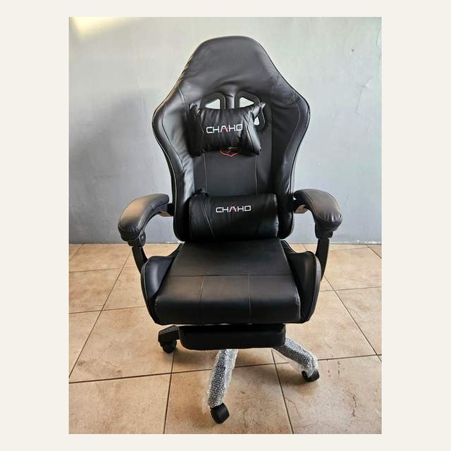Kursi gaming hitam tampak depan dengan dua bantal bertuliskan CHAHO dan kaki beroda terbungkus sebagian.