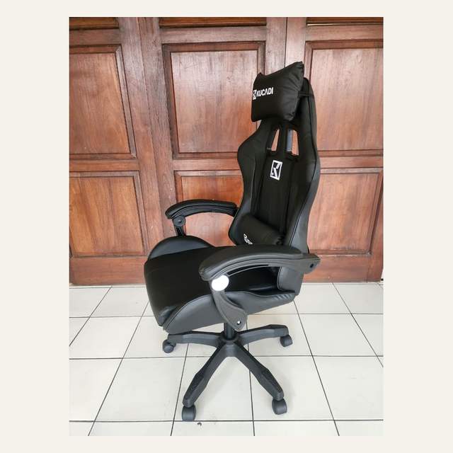 Kursi gaming hitam tampak serong dengan bantal kepala bertuliskan KUCADI dan kaki lima cabang beroda.