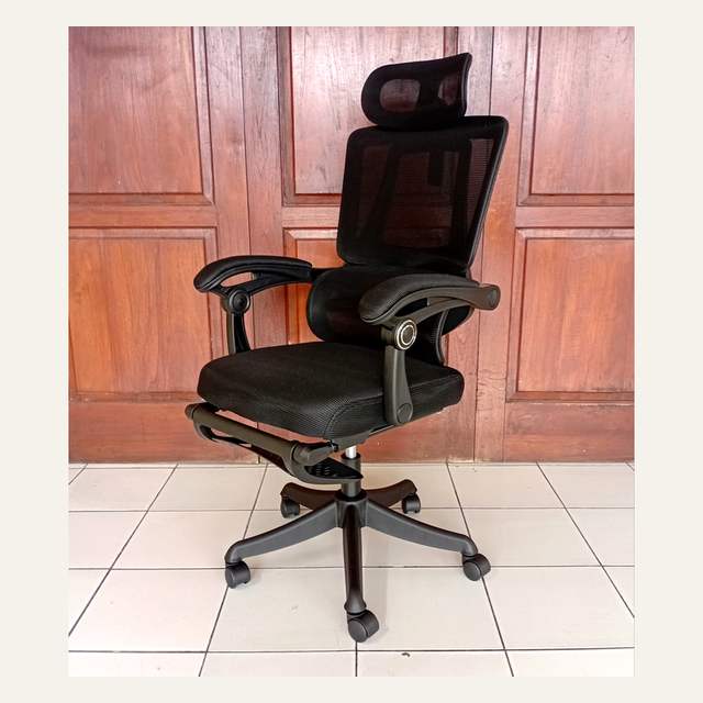 Kursi kantor hitam tampak serong dengan sandaran jaring dan sandaran kaki terlipat di bawah dudukan.