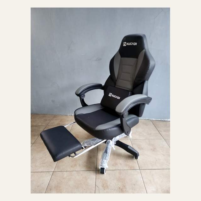 Kursi kerja hitam abu-abu bertuliskan KUCADI dengan sandaran kaki terbuka, difoto serong di depan dinding abu-abu.