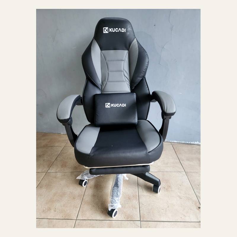 Kursi hitam abu-abu bertuliskan KUCADI difoto dari depan dengan bantal kecil di dudukan.