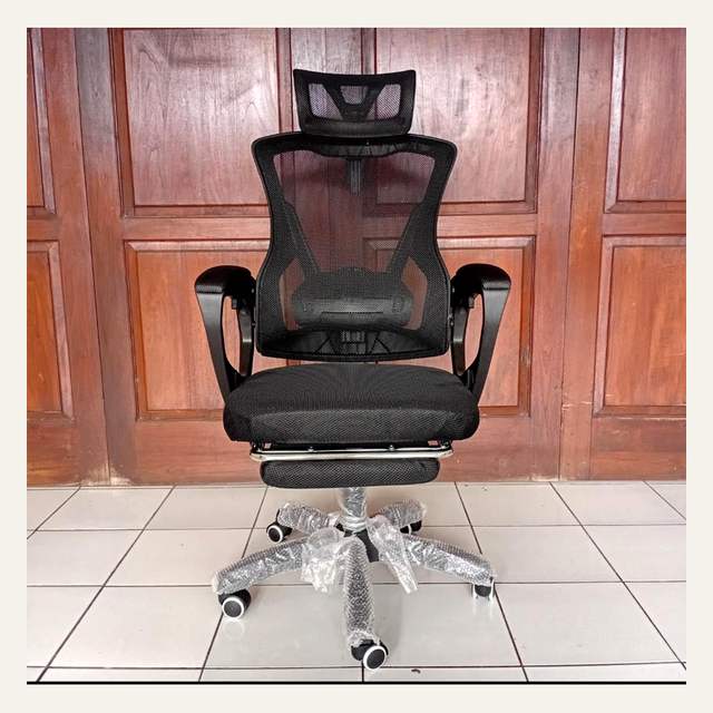 Kursi kantor hitam bersandaran jaring dengan sandaran kepala dan sandaran kaki di depan panel kayu.