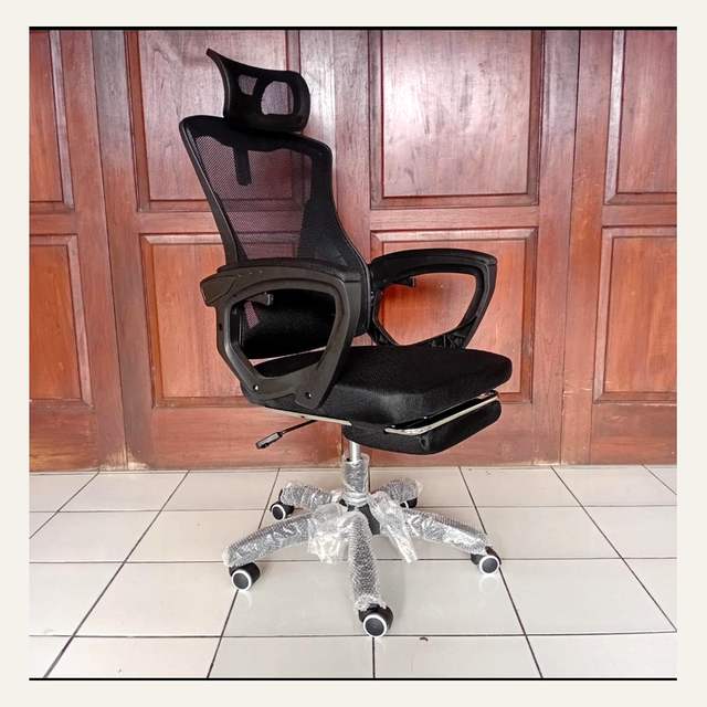 Kursi kantor hitam bersandaran jaring difoto dari samping di depan panel kayu cokelat.