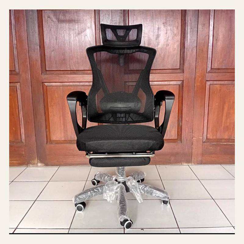 Kursi kantor hitam bersandaran jaring dengan sandaran kepala dan sandaran kaki di depan panel kayu.