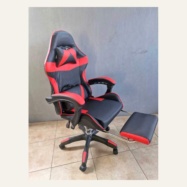 Kursi hitam merah dengan bantal leher dan sandaran kaki terbuka difoto dari sudut serong.