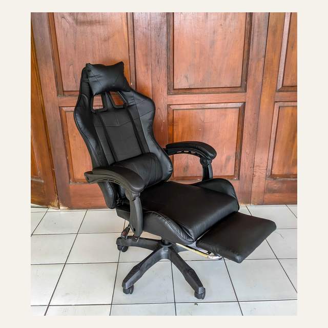 Kursi hitam polos dengan sandaran kaki terbuka penuh, difoto serong di depan pintu kayu.