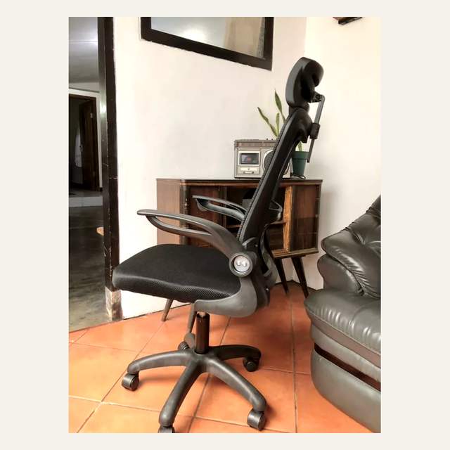 Kursi kantor hitam difoto dari samping di ruangan berlantai oranye dengan lemari kayu di belakang.