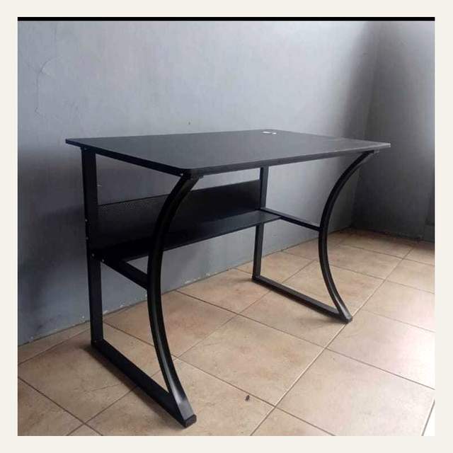 Meja hitam berkaki melengkung dengan rak bawah dan lubang kabel, foto agak buram beresolusi rendah.