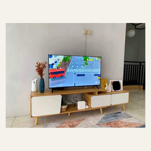 Rak tv kayu terang berpanel putih dengan televisi menyala dan vas di atasnya di ruang berdinding putih.