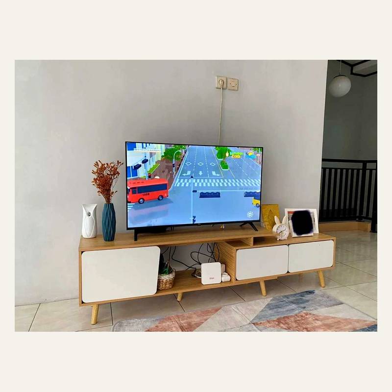 Rak tv kayu terang berpanel putih dengan televisi menyala dan vas di atasnya di ruang berdinding putih.